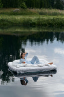 A woman relaxes on a white mattress floating on calm water, with a grassy shoreline and trees in the background. Her reflection is visible in the water.